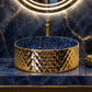 A luxury bathroom vanity setup featuring a round gold vessel sink with a textured diamond pattern, set into a blue marble countertop and matching backsplash. A large illuminated LED ring mirror hangs above, with decorative gold and black vases on either side.