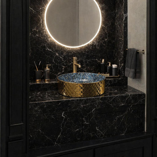 A luxury bathroom vanity featuring a black marble countertop and matching marble backsplash with a glowing circular LED mirror. A gold-finished vessel sink with a blue marbled interior sits on the counter, flanked by gold fixtures.