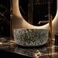 A round vessel sink with a black, white, and gold marbled pattern sits on a black marble countertop with gold veining, paired with a brushed gold faucet in a luxurious bathroom setting.