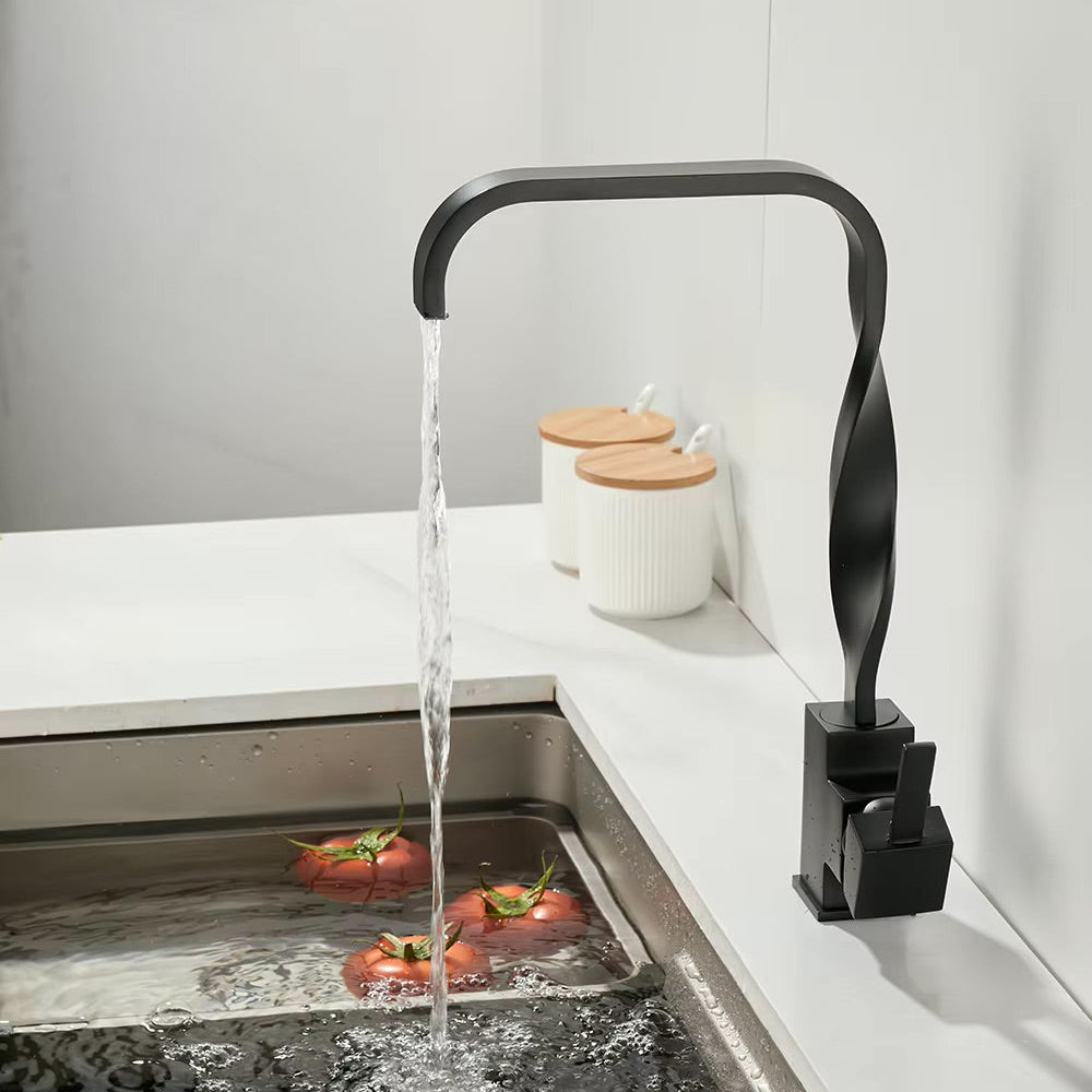 A modern matte black twisted gooseneck faucet running with clear water into a stainless steel sink, with tomatoes washing in the water and two white canisters with wooden lids in the background.