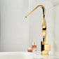 A modern high-arc gold faucet with a polished chrome base, shown installed on a white sink with cosmetic bottles in the background.
