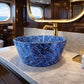 A blue marbled ceramic vessel sink with gold flecks, set into a white marble bathroom vanity countertop with a gold faucet, on a yacht with an ocean view visible through the window.