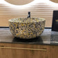 A round vessel sink with a black, gold, and yellow marbled pattern, set on a dark granite countertop with a gold pull handle. A black card with gold script branding is visible on the right.