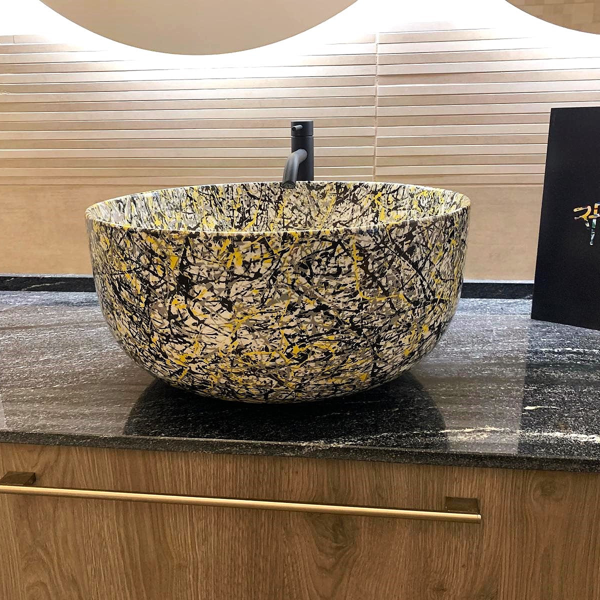 A round vessel sink with a black, gold, and yellow marbled pattern, set on a dark granite countertop with a gold pull handle. A black card with gold script branding is visible on the right.